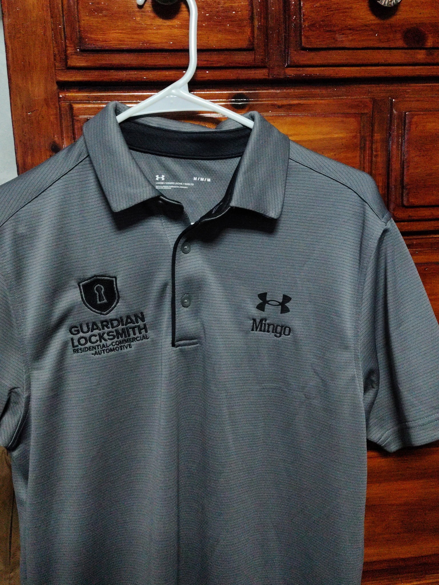 Gray polo shirt with "Guardian Locksmith" logo and "Mingo" embroidered, hanging on a white plastic hanger against wooden background.